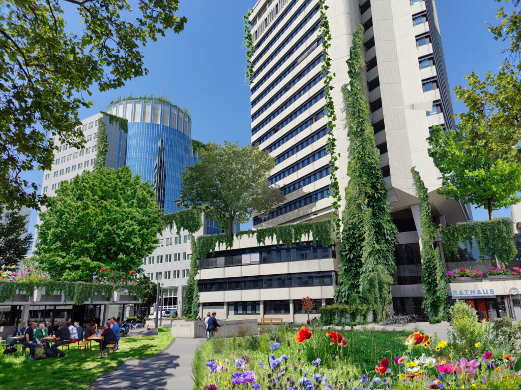 Begrünte Gebäude rund um den Stadthof, mit Blumenwiese und Tische und Bänken mit Leuten